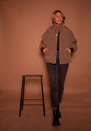 Manteau mouton à col montant porté par une femme, silhouette entière, tabouret noir, jean noir, de face