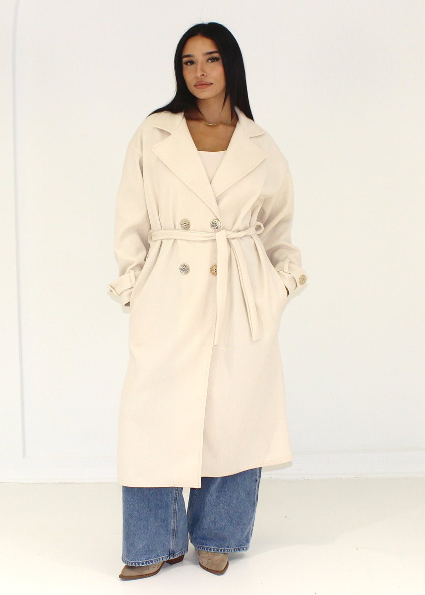 Manteau beige à boutons dorés — vue de face, ceinture nouée.