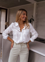 Chemise brodée à manches longues portée par une femme, vue de trois-quarts, mains sur les hanches, pantalon beige