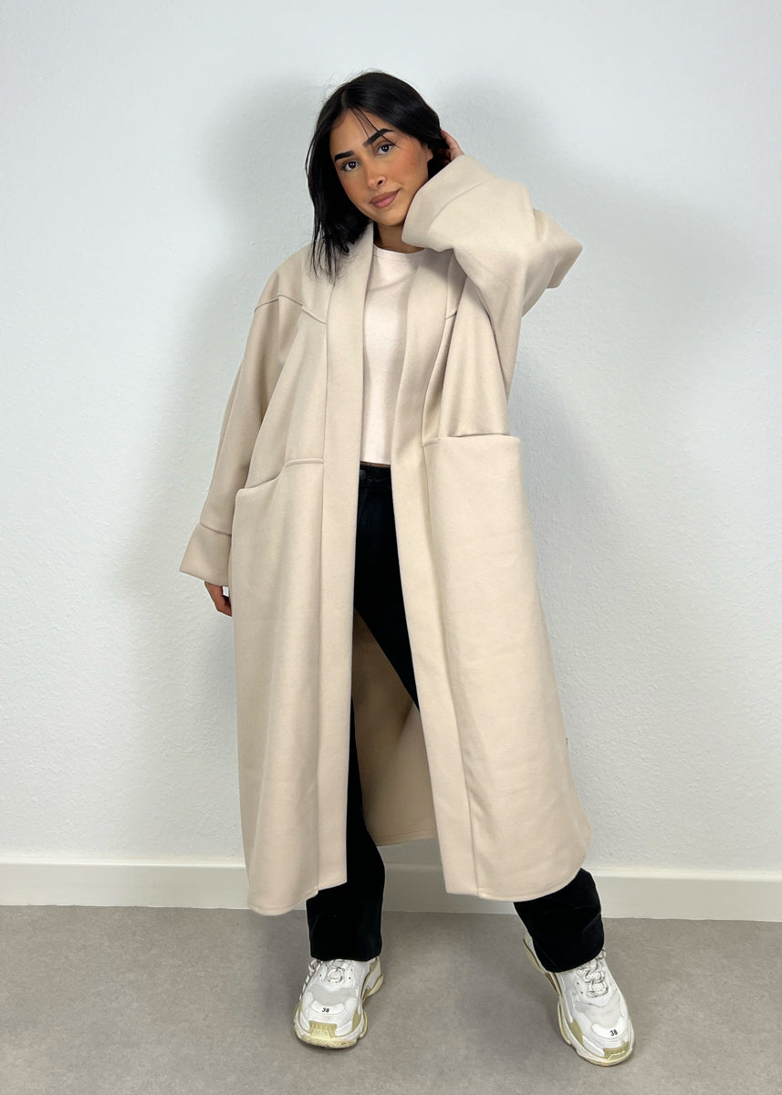 Manteau oversize beige — vue portée de face, main sur la hanche, pantalon clair et baskets blanches