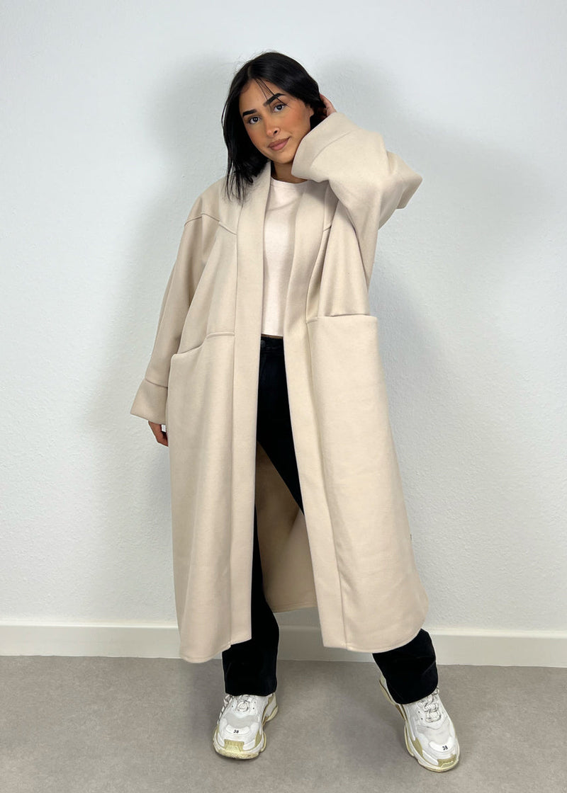 Manteau oversize beige — vue portée de face, main sur la hanche, pantalon clair et baskets blanches