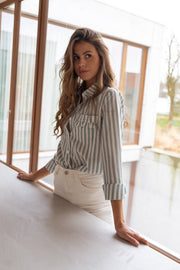 Chemise rayée à poches volantées portée par une femme, silhouette entière, baie vitrée, pantalon beige