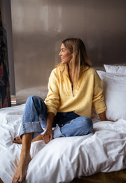 Pull loose col camionneur porté par une femme, assise, sur un lit, jean