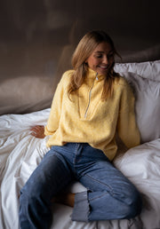 Pull loose col camionneur porté par une femme, assise, sur un lit blanc, jean