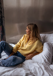 Pull loose col camionneur porté par une femme, assise, sur le lit, regard vers la fenêtre, de côté