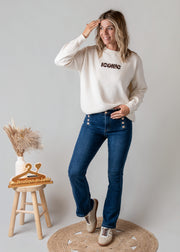 Jean flare à boutons porté par une femme, silhouette entière, pull blanc, tabouret