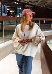 Sweat doux à écritures porté par une femme, vue de trois-quarts, casquette rose, jean, baie vitrée