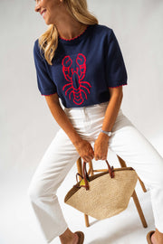 Pull en maille manches courtes porté par une femme, vue de trois-quarts, sac en paille, pantalon blanc