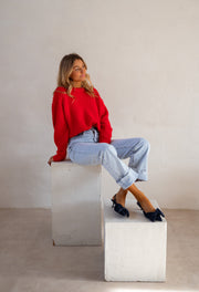 Pull col rond porté par une femme, assise, sur un cube blanc, escarpins noirs, de loin