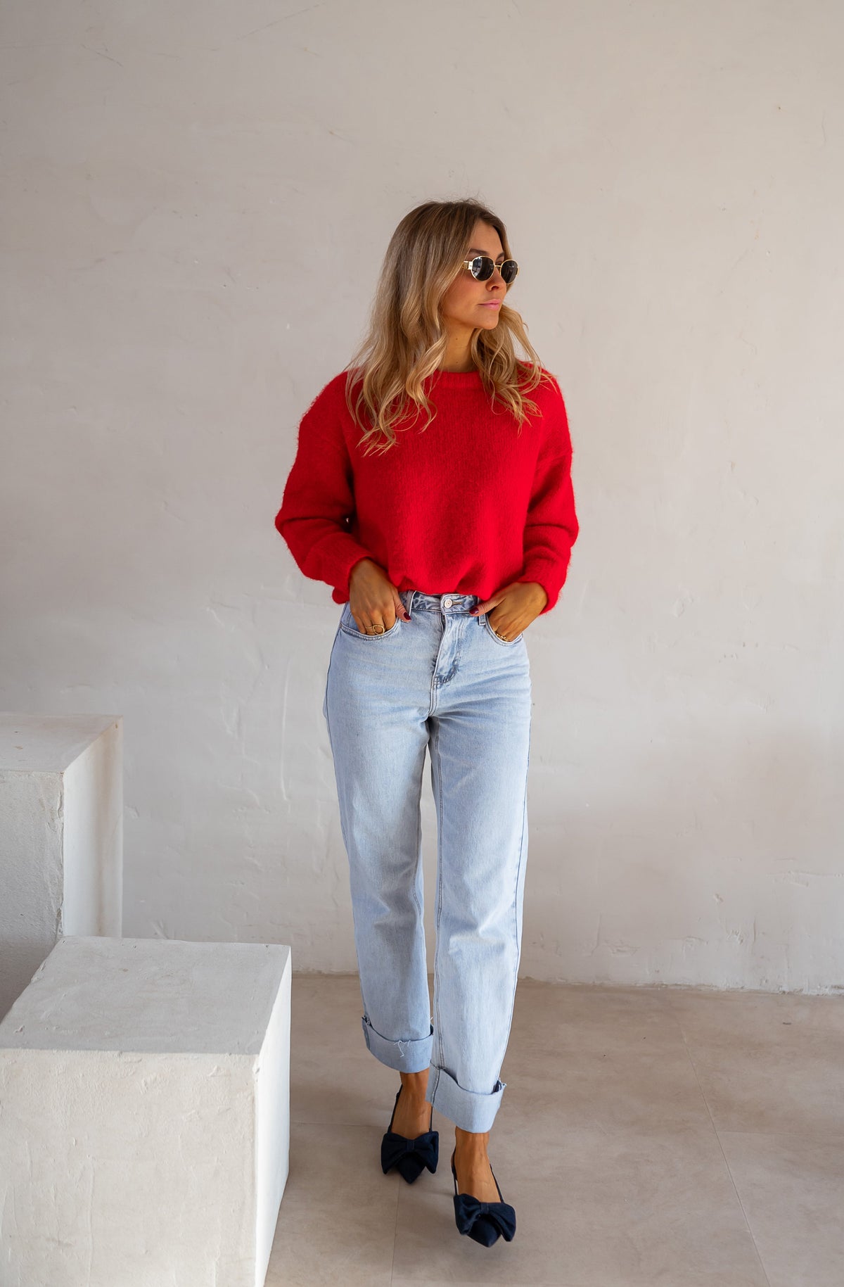 Pull col rond porté par une femme, silhouette entière, jean clair, escarpins noirs, mur blanc