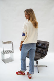 Pull à logo manches longues écru porté par une femme, vue de dos, fauteuil noir, jean bleu