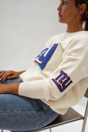 Pull à logo manches longues écru porté par une femme, assise, fauteuil noir, logo sur la manche
