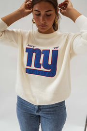 Pull à logo manches longues écru porté par une femme, vue rapprochée, logo bleu, mains dans les cheveux