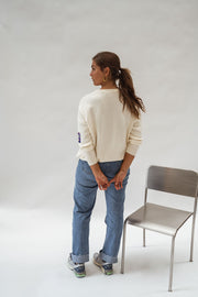 Pull à logo manches longues écru porté par une femme, vue de dos, chaise grise, queue-de-cheval