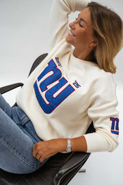 Pull à logo manches longues écru porté par une femme, vue rapprochée, logo bleu, fauteuil noir