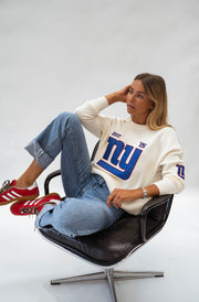 Pull à logo manches longues écru porté par une femme, assise, fauteuil noir, baskets rouges