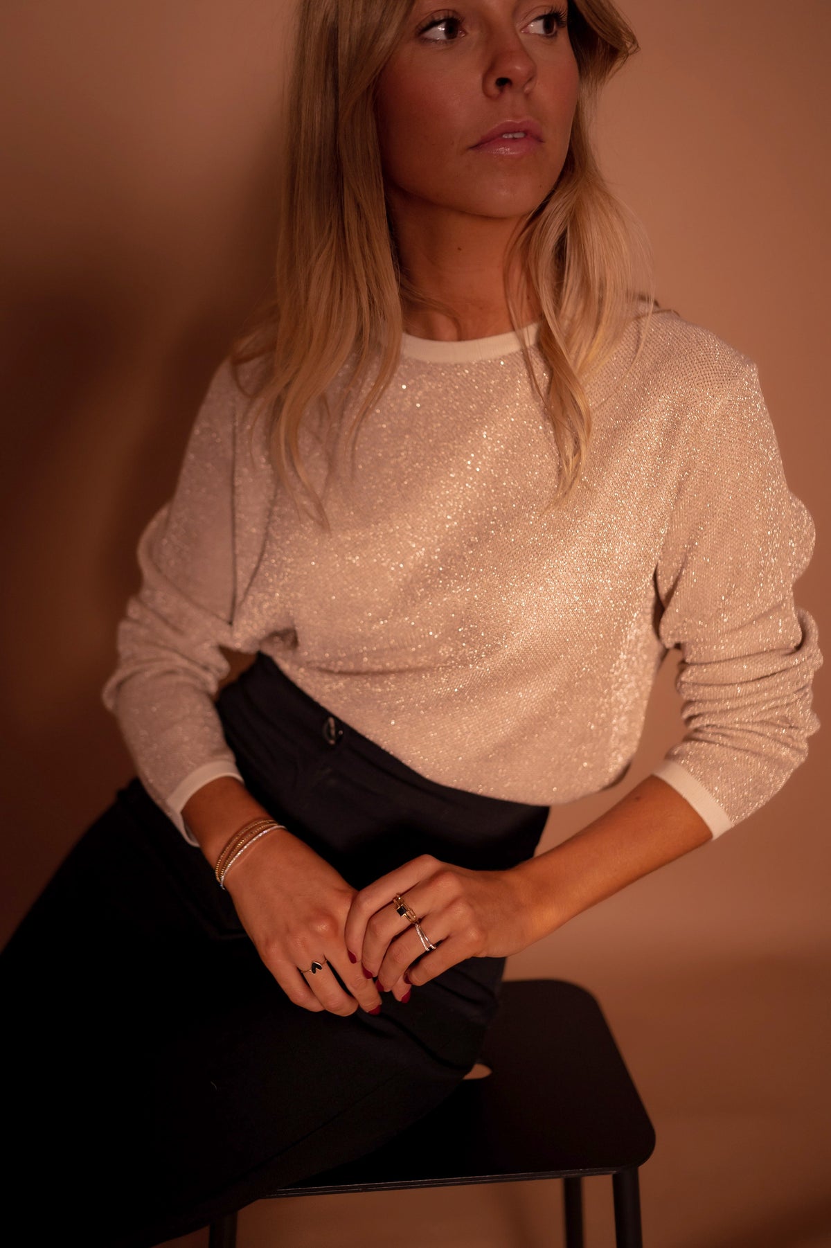 Pull à paillettes col rond porté par une femme, vue rapprochée, pantalon noir, mains jointes