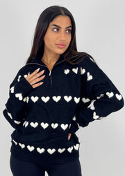 Pull à fermeture zippée noir — vue portée de face, cadrée à la taille, col entrouvert, petits cœurs blancs
