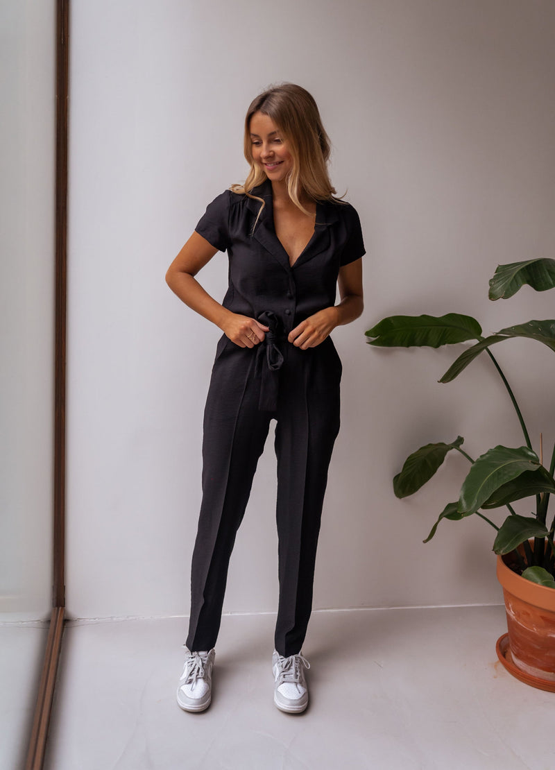 Combinaison fluide à col chemise portée par une femme, vue de face, mur blanc, plante verte