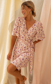 Robe fleurie cache-cœur courte portée par une femme, vue de trois-quarts, rideaux, jambe pliée