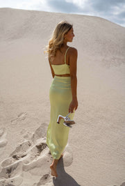 Robe longue effet soie verte portée par une femme, vue de dos, dunes, sandales à la main