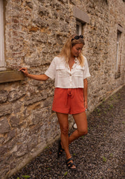 Short court à taille élastique porté par une femme, silhouette entière, mur en pierre, chemise blanche