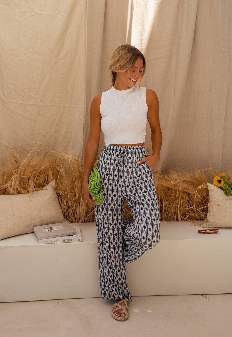 Pantalon fluide à motif tie-dye porté par une femme, silhouette entière, rideau, sac vert