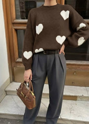 Pull à motif cœurs — vue portée de trois-quarts, en extérieur devant une façade, pantalon plissé gris foncé et petit sac