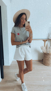 T-shirt à motif cœur porté par une femme, silhouette entière, chapeau, short blanc, baskets