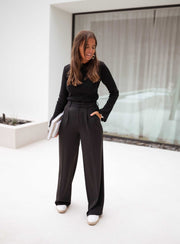Pantalon à pinces noir porté par une femme, vue de trois-quarts, haut noir, mains sur les hanches