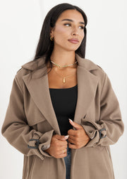 Trench oversize taupe à ceinture — vue portée de face, cadrée à la taille, regard sur le côté