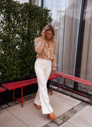 Chemisier fluide à motifs dorés porté par une femme, silhouette entière, pantalon blanc, banc rouge